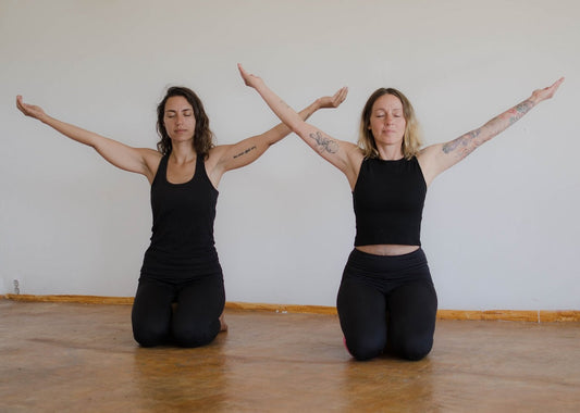 Katonah-Yoga: Zwei Frauen sitzen im Fersensitz und strecken beide Arme zu den Seiten und nach oben