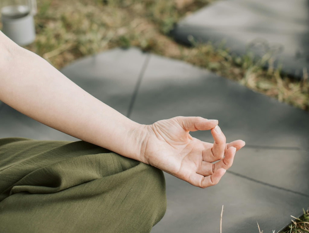 Hand im Meditations-Mudra auf dem Knie abgelegt, Yogamatte und Wiese im Hintergrund 